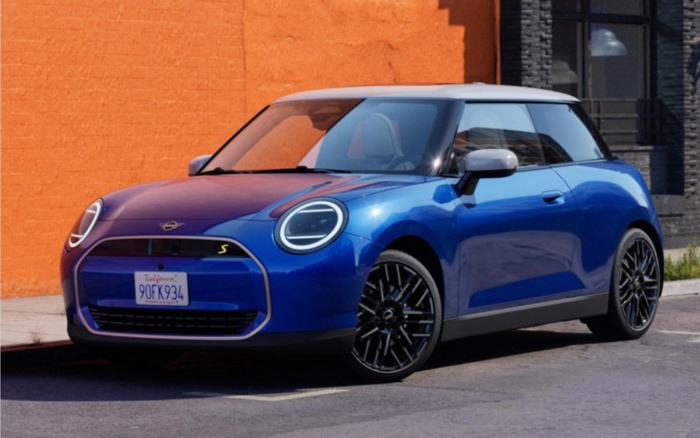 Blue Mini Cooper SE electric hatchback car with white roof parked on street near orange and grey buildings, featuring modern circular LED headlights and California license plate 90FK934, representing new cars and used cars options.