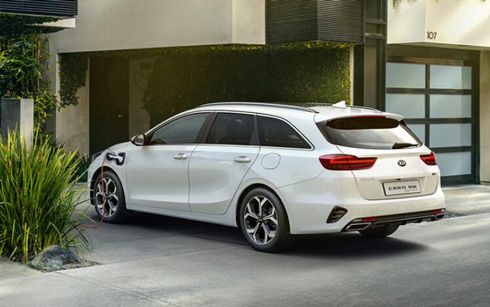 White Kia Ceed SW plug-in hybrid car parked outside a modern house at 107, charging with an electric cable connected to the front left side. The car is a used or new car option, showcasing eco-friendly technology in a residential setting.