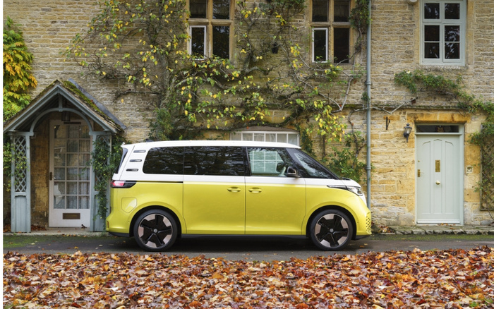 Yellow and white modern electric minivan parked on a street next to a stone cottage with greenery climbing the walls and doors, fallen autumn leaves covering the ground, highlighting a beautiful example of new cars available alongside used cars.