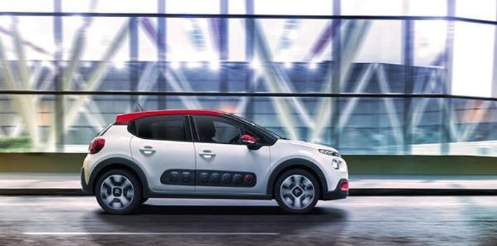 Side view of a white and red compact car driving on a road with a modern glass building in the background, showcasing a stylish used car option from a dealership offering both used cars and new cars.