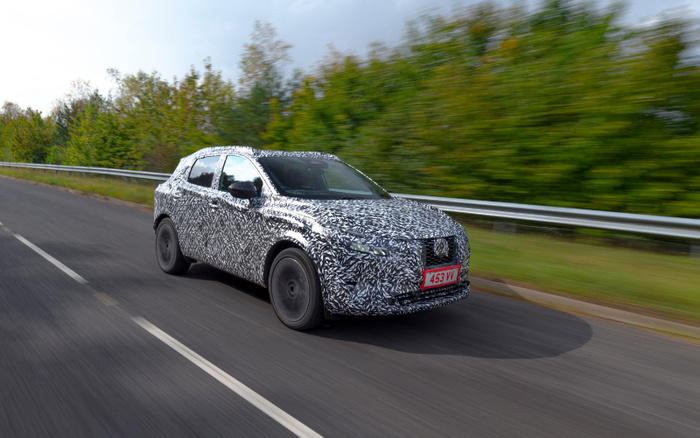 A camouflaged test car driving on a road lined with green trees and metal guardrails, featuring a black and white swirl pattern to conceal its design details. The car has a red license plate with the registration number 453 IV. This image represents new cars in development, relevant to buyers interested in both used cars and new cars.