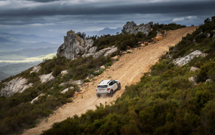 White off-road vehicle driving up a rugged dirt path on a mountainous terrain with rocky outcrops and dense green shrubs under a cloudy sky, showcasing the capability of new cars and used cars to handle rough landscapes.