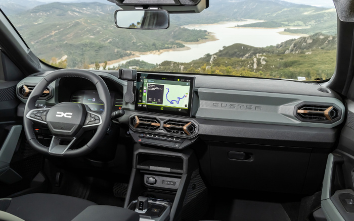 Interior dashboard and steering wheel of a Dacia Duster car, featuring a digital instrument cluster, touchscreen navigation display showing a map, and hexagonal air vents. The view through the windshield reveals a scenic landscape of hills and a winding river. Ideal for browsing used cars or new cars with modern technology and rugged design.