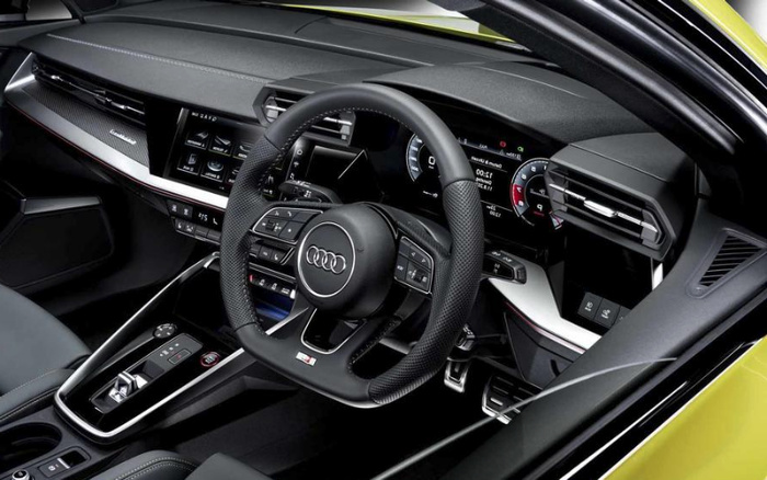 Interior view of a new Audi car showing the steering wheel with the Audi logo, digital instrument cluster displaying speed and navigation, central touchscreen with various controls, and gear selector area. The modern dashboard features sleek design elements and the word 'quattro' on the trim, highlighting features found in new cars and used cars.