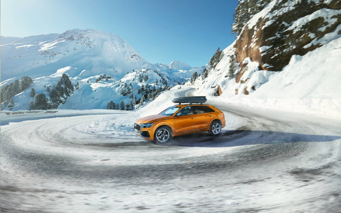 Orange SUV driving on a snowy mountain road with skid marks on the snow, surrounded by snow-covered trees and mountains under clear blue sky, showcasing a new car in a winter landscape, suitable for promoting used cars and new cars capable of handling snowy conditions.