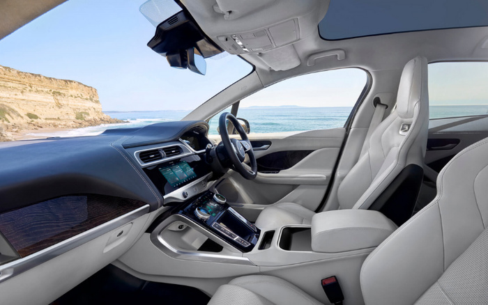Interior of a modern car showing the driver's seat, steering wheel, and dashboard with digital displays, featuring light grey leather seats and black and wood grain dashboard accents, with a coastal cliff and ocean visible through the windows, representing used cars and new cars.