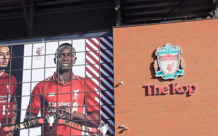 Exterior view of The Kop stand at Liverpool Football Club stadium featuring a large mural of Liverpool FC players wearing red kits with the Liverpool FC crest displayed prominently alongside the text 'The Kop' on a brick wall