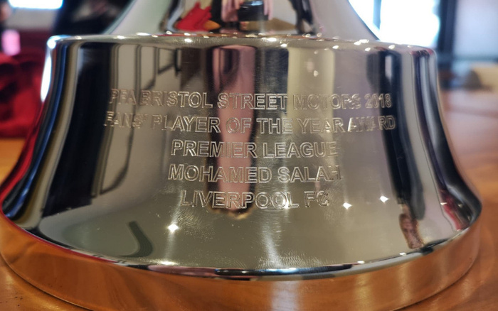 Close-up of a shiny silver trophy engraved with the text 'PFA Bristol Street Motors 2018 Fans' Player of the Year Award Premier League Mohamed Salah Liverpool FC' reflecting its surroundings. The image highlights the trophy awarded to Mohamed Salah by Bristol Street Motors, a company known for used cars and new cars sales.