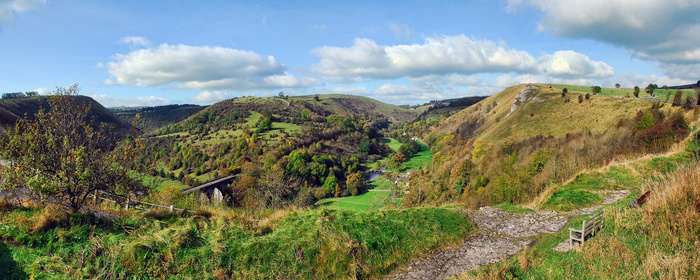 Panoramic view of a green valley with grassy hills, trees, a bridge, and a walking path with a wooden bench on the right under a partly cloudy blue sky. No text or cars visible in the natural landscape, not depicting used cars or new cars.
