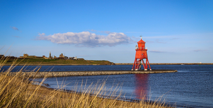 Red lighthouse on a pier with calm water and a clear blue sky, with a grassy shoreline and distant buildings under a cloud, no text or references to used cars or new cars