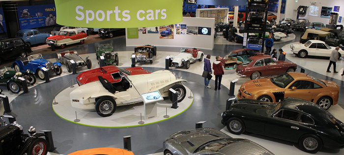 Interior of a car museum showcasing a variety of vintage and classic sports cars displayed in a circular arrangement under a large green sign reading 'Sports cars'. The exhibition includes various models in different colours such as white, red, blue, green, and orange, with visitors walking and observing the cars. The setting highlights both used cars in historic condition and new cars inspired by classic designs.