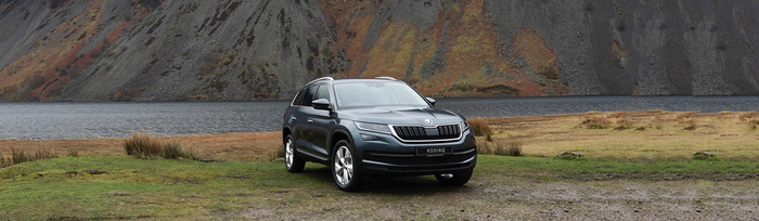 Grey Škoda Kodiaq SUV parked on grassy terrain near a lake with steep rocky hills in the background. The car's front license plate reads 'KODIAQ'. Scenic outdoor setting highlighting the vehicle among nature, suitable for used cars and new cars listings.