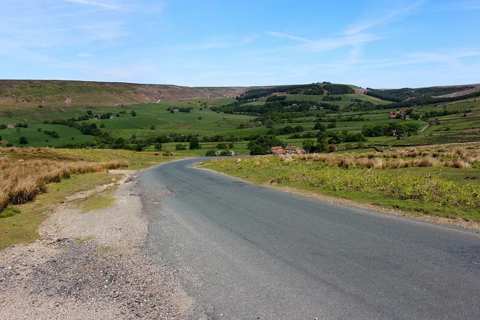 Empty winding road through a green countryside landscape with rolling hills and scattered houses under a clear blue sky, no used cars or new cars visible in the image.