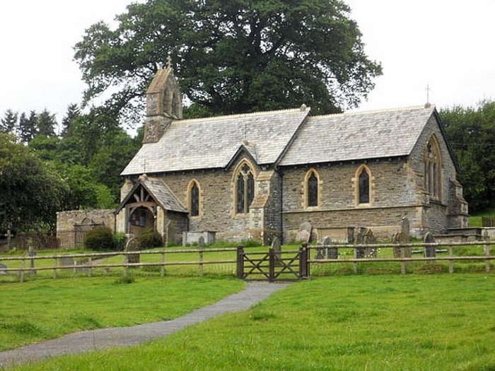 Stone church building with a slate roof surrounded by a grassy graveyard, featuring a wooden gated fence and trees in the background. No text visible in the image. Keywords: used cars, new cars.