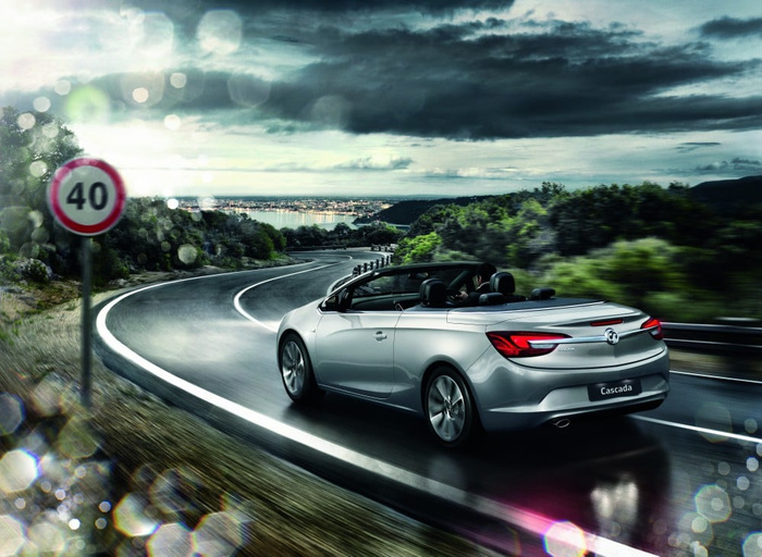Silver Vauxhall Cascada convertible car driving on a winding road near a coastal area under a dramatic cloudy sky with a 40 mph speed limit sign visible on the left. The image highlights the sleek design of the Cascada, suitable for both used cars and new cars buyers looking for stylish convertibles.