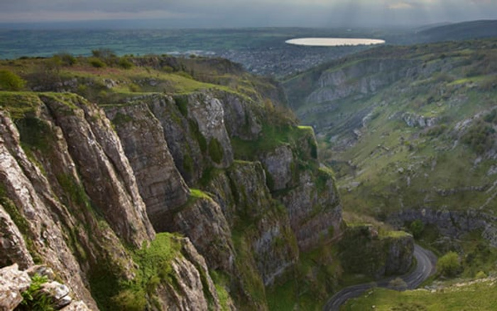 A scenic view of a rugged green valley with steep rocky cliffs and a winding road below, overlooking a distant town with a large lake, suitable for background images for used cars and new cars advertisements.