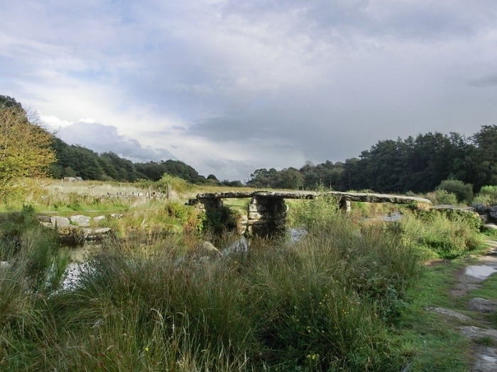 Stone clapper bridge over a small river in a rural countryside setting with grassy banks and trees under a cloudy sky, no text, no cars visible