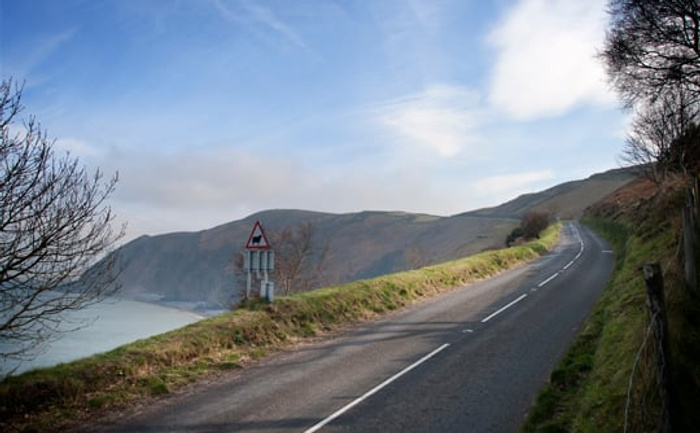 Winding coastal road with clear lane markings, flanked by grassy slopes and trees, overlooking a large body of water and distant hills under a partly cloudy sky. No vehicles are present, symbolising a scenic drive suitable for used cars and new cars.