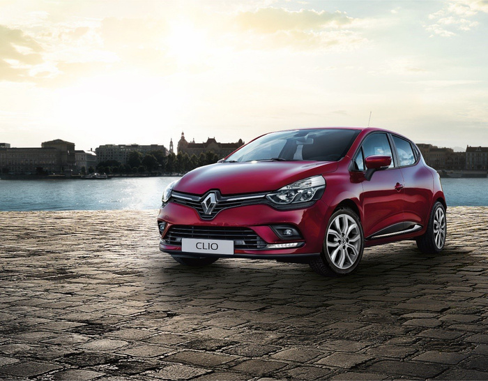 Red Renault Clio parked on a cobblestone pavement near a waterfront with city buildings in the background during sunset, showcasing a new car model.