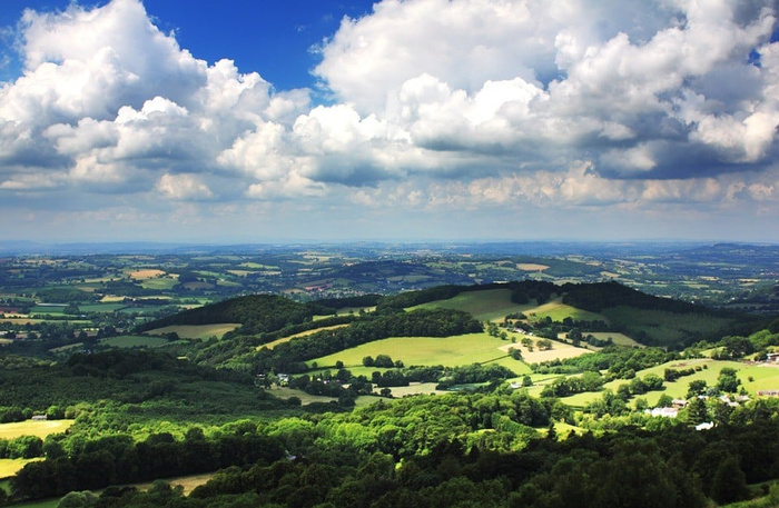 Scenic countryside landscape with lush green fields, rolling hills, scattered trees, and a partly cloudy blue sky stretching over the horizon. No text or cars visible in the image.