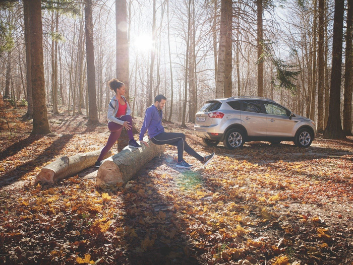 A silver SUV parked in a sunlit forest clearing during autumn, with two people exercising on large tree logs covered by fallen leaves. The scene highlights outdoor activity and nature alongside the vehicle, representing both new cars and used cars in a natural setting.