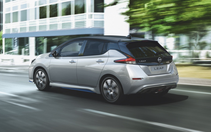 Silver Nissan Leaf electric car driving on a city road, featuring black roof and rear design details. The car's model name 'LEAF' is displayed on both the back and the license plate. Background shows modern glass buildings and green trees, representing a clean urban environment ideal for both new cars and used cars.