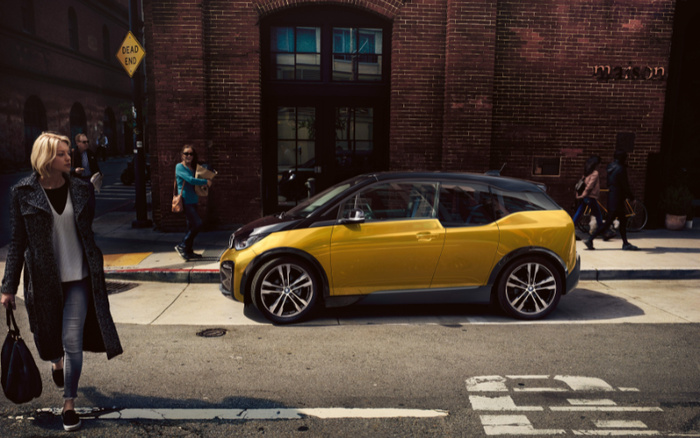 Yellow and black compact car parked on a city street in front of a red brick building with a sign partially visible reading 'maison'. Pedestrians walking nearby, including a blonde woman in a dark coat crossing the street holding a black handbag. A yellow diamond Dead End road sign is visible on a brick wall. The image features a modern used car or new car in an urban setting.
