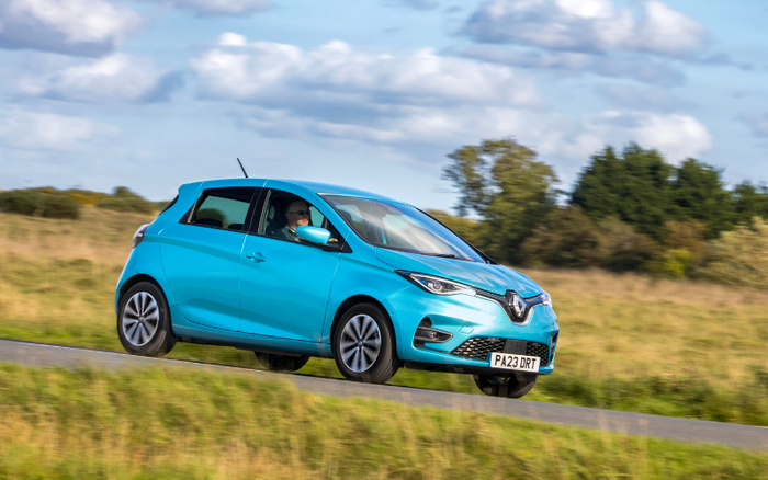 Bright blue Renault Zoe electric hatchback driving on a country road with a woman wearing sunglasses behind the wheel. The car has a UK registration plate PA23 DRT. The background features green grass, trees, and a partly cloudy sky. Suitable for used cars and new cars listings.