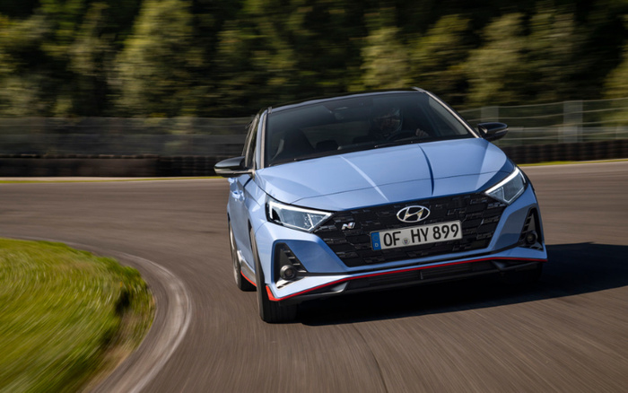 Blue Hyundai i20 N driving on a curved race track with green trees in the background, showcasing a modern front design with distinctive headlights and a license plate reading OF HY 899, representing new cars and used cars available for sale.