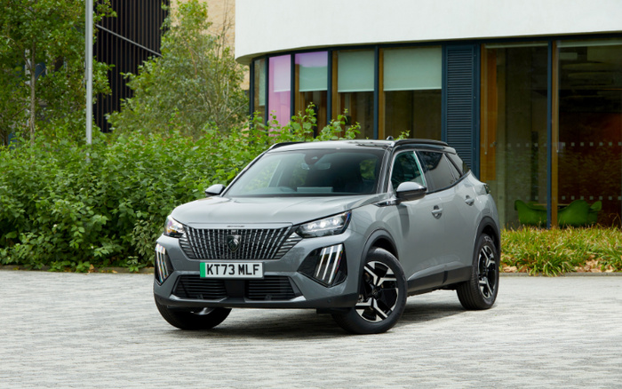 Grey Peugeot SUV with registration plate KT73 MLF parked on a paved area in front of a modern building with large windows and greenery in the background, suitable for advertising used cars and new cars.