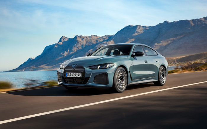 A sleek metallic blue BMW sedan driving on a coastal road with mountains in the background, showcasing a modern design with distinctive kidney grille and LED headlights, ideal for those interested in used cars and new cars.