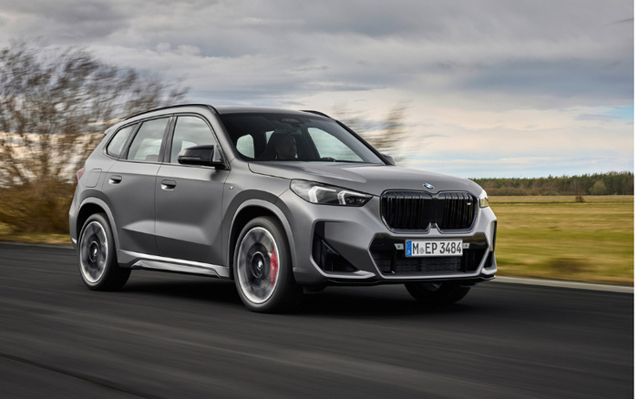 Grey BMW SUV driving on a road with blurred trees and overcast sky in the background, featuring German license plate M EP 3484, representing used cars and new cars.