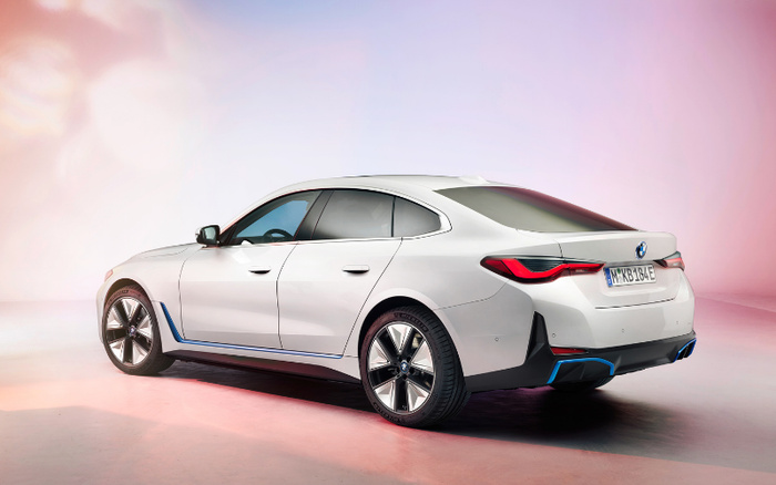 Rear and side view of a white BMW i4 electric car displayed in a studio with pink and white lighting, highlighting its sleek design. The vehicle has distinctive blue accents and modern alloy wheels, suitable for advertising used cars and new cars.