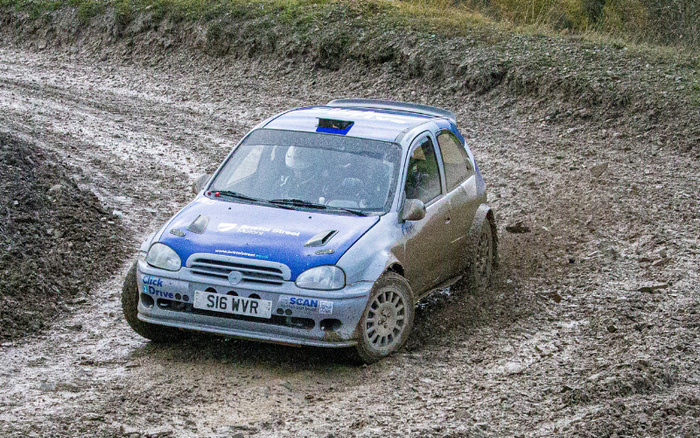 Blue and white rally car with registration plate S16 WVR driving on a muddy dirt track, showing sponsors including Click2Drive, Scan, and Total Direct Energy on the front and hood, in a rural outdoor setting with visible tyre tracks on the ground, representing used cars and new cars in a rugged environment.