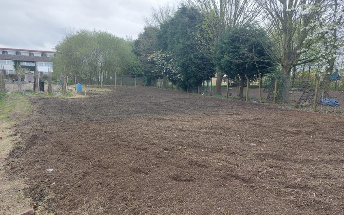 A freshly tilled open plot of land with soil turned over, bordered by a wooden fence and trees on the right side, with residential buildings partially visible in the background under a cloudy sky. No cars or related text are visible in the image, so it does not depict used cars or new cars.