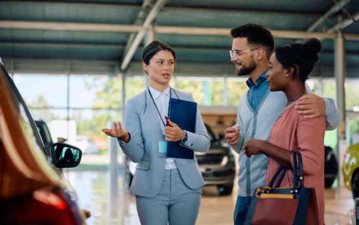 Car dealership salesperson in a grey suit holding a blue folder talking to a smiling couple inside a showroom with new cars, discussing options. The man has his arm around the woman, and the setting suggests a professional consultation about used cars and new cars available for purchase.
