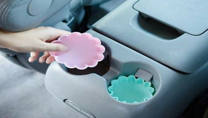 Close-up of a car's centre console cup holder with a light pink flower-shaped silicone coaster being placed inside by a hand, and a light blue flower-shaped silicone coaster already in the adjacent cup holder, highlighting accessories suitable for used cars and new cars.