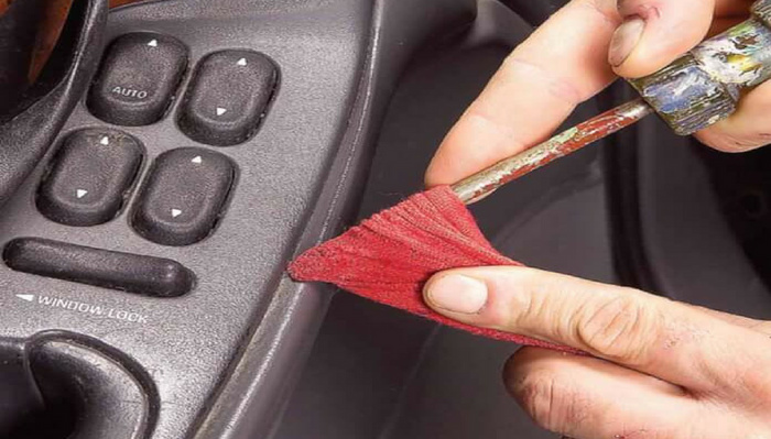 Close-up of a car's power window control panel featuring four black buttons with white arrows and the label 'WINDOW LOCK'. A hand is using a small red cloth and a rusty metal tool to clean the panel, highlighting maintenance or restoration of used cars and new cars.