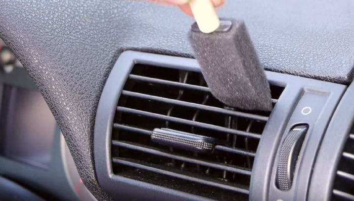 Close-up of a car's air vent on the dashboard with a person cleaning the vent slats using a small foam brush. The vent features a textured adjustment knob and a proximity switch with on and off settings. This image represents maintenance and care important for both used cars and new cars.