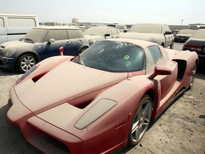 Dust-covered red Ferrari Enzo sports car parked in an outdoor lot with several other dusty cars including a Mini Cooper and SUVs in the background, indicating a storage or holding area for used cars and new cars.
