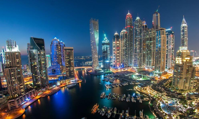 Nighttime panoramic view of a city marina with illuminated skyscrapers, modern high-rise buildings, and reflections of city lights on the water. Numerous boats and yachts are docked along the marina. This vibrant urban scene represents a modern cityscape but does not feature used cars or new cars.