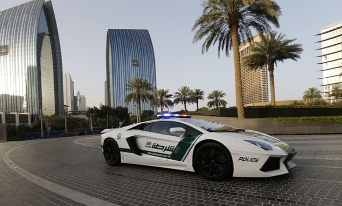 White and green Dubai police Lamborghini sports car with emergency lights on, parked on a cobblestone street in a modern cityscape featuring tall curved glass skyscrapers and palm trees. The car has Arabic text and 'POLICE' written on the side. Urban setting with clear sky, highlighting an example of new cars rather than used cars.