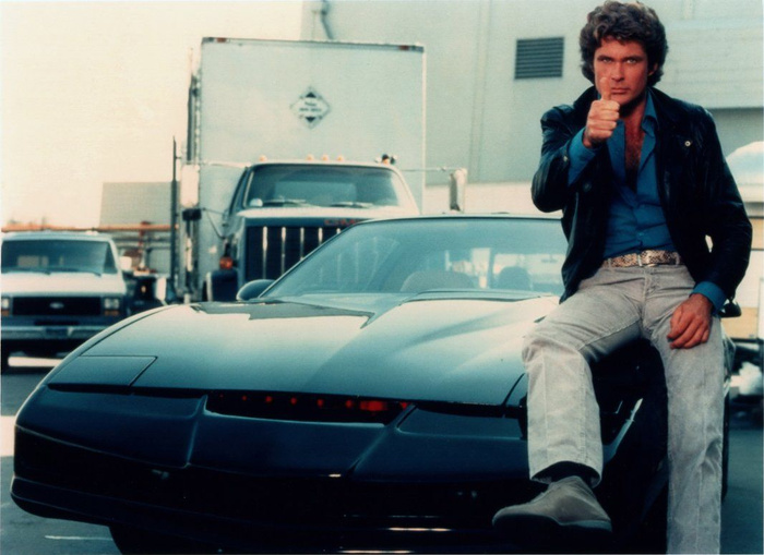 Man in a black leather jacket and blue shirt giving a thumbs up while sitting on the hood of a sleek black sports car resembling the Knight Rider car, parked in an industrial area with trucks in the background, representing themes related to used cars and new cars.