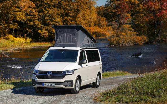 White Volkswagen camper van with a pop-up roof parked on a dirt path beside a river with autumn trees in the background, showcasing a scenic outdoor setting, ideal for advertisements featuring used cars and new cars.