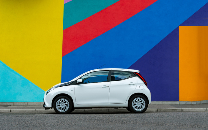 White compact car parked on the street in front of a colourful wall with geometric shapes in yellow, turquoise, red, blue, purple, and orange. The image highlights a modern, small hatchback suitable for city driving, representing options for both used cars and new cars.