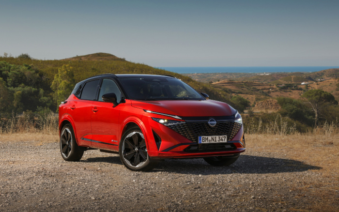 Red Nissan SUV parked on a gravel area overlooking a scenic landscape with hills and the sea in the background, showcasing a sleek modern design. The car is a new model with black roof and alloy wheels, suitable for buyers interested in used cars or new cars.