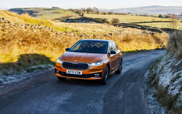 Orange Skoda Fabia driving on a narrow country road with grassy and rocky hills in the background, registration plate KY71 YSW, suitable for advertisements about new cars and used cars.