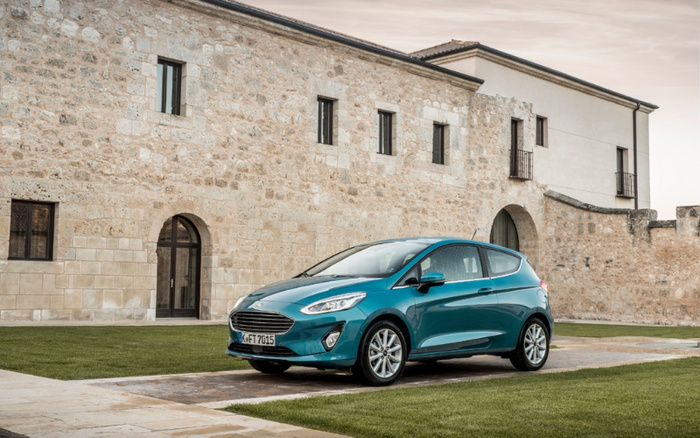 Blue Ford Fiesta hatchback car parked on a paved driveway next to a historic stone building with multiple arched doors and small windows, illustrating a modern new car model suitable for used cars and new cars buyers.