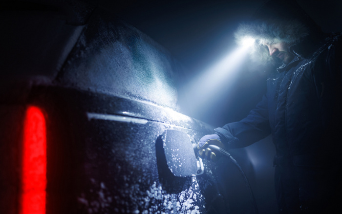 Person wearing winter clothing with fur hood and gloves charging an electric car at night, with the car covered in frost and ice, illuminated by a bright headlamp and the red rear light of the vehicle, highlighting the cold weather conditions for used cars and new cars.