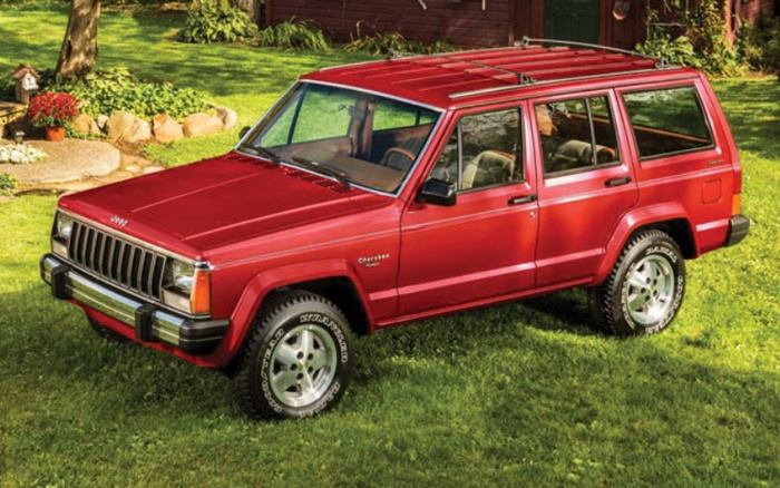 Red Jeep Cherokee Sport parked on green grass in front of a wooden shed, showcasing a classic 4x4 SUV with visible Jeep logo on the front grille and 'Cherokee Sport' badge on the side, representing a used car model against a natural garden background, suitable for new cars and used cars advertisements.